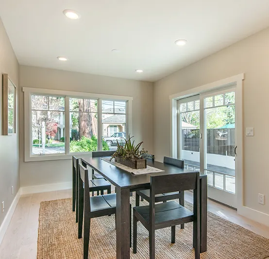 A modern dining room with a large wooden table, black chairs, and a jute rug. The room has large windows with sliding glass doors leading to a patio.