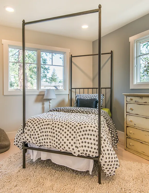 A bedroom with a metal four-poster bed, a polka-dot duvet, and a vintage dresser. The room has light-colored walls, a large window, and a fluffy rug on the floor.