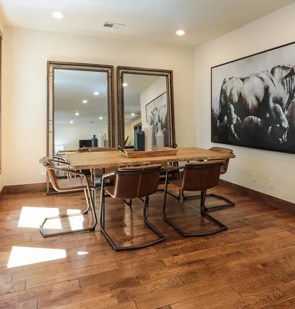 A modern dining room with a wooden table, leather chairs, and a large black and white photograph of a cow on the wall. The room features two large mirrors and hardwood floors