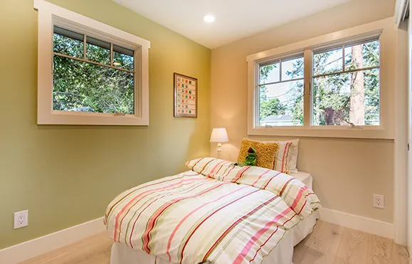 A cozy bedroom with a single bed, a striped duvet, and a framed poster on the wall. The room has light green walls, a wooden floor, and large windows overlooking a backyard