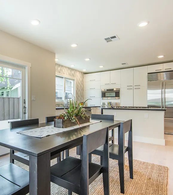 A modern kitchen and dining room combined in an open-plan space. The kitchen features white cabinets, stainless steel appliances, and a large island. The dining area has a wooden table and black chairs.