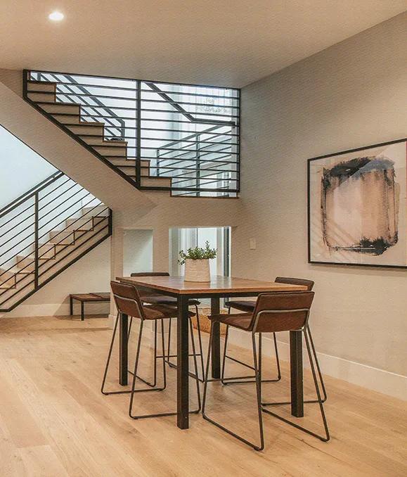 A modern dining room with a wooden table, metal chairs, and a metal staircase leading to an upper level. The room has light-colored walls and a hardwood floor