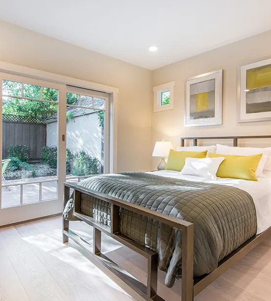 A modern bedroom with a king-sized bed, white bedding, and yellow throw pillows. The room has light-colored walls, a wooden floor, and large sliding glass doors leading to a patio