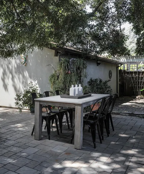 A modern outdoor patio with a large rectangular table, metal chairs, and potted plants. The patio is shaded by a large tree and surrounded by greener