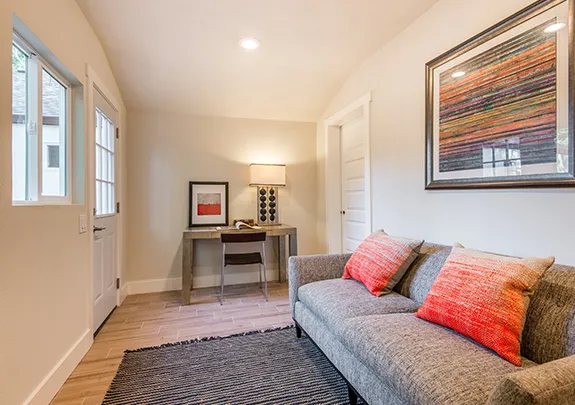 A multi-functional room with a gray sofa, orange pillows, a small desk, and a large abstract painting on the wall. The room has a light-colored ceiling, white walls, and a wooden floor