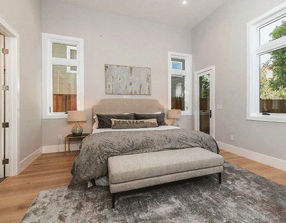 A modern bedroom with a king-sized bed, gray bedding, and a neutral color palette. The room features large windows, a wooden floor, and a bench at the foot of the bed