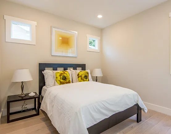 A modern bedroom with a queen-sized bed, white bedding, and yellow throw pillows. The room has light-colored walls, a wooden floor, and a framed abstract painting above the bed