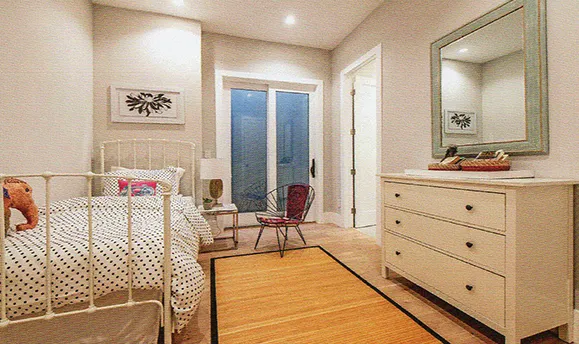 A cozy bedroom with a white metal bed, a patterned duvet, and a plush elephant toy. The room has light-colored walls, a wooden floor, and a large sliding glass door leading to a patio