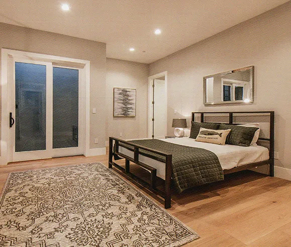 A modern bedroom with a metal bed frame, a patterned duvet, and a rug on the floor. The room has light-colored walls, a large sliding glass door, and a ceiling fan