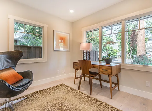 A modern home office with a mid-century modern desk, a leather armchair, a patterned rug, and large windows overlooking a backyard.