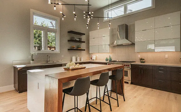 A modern kitchen with white cabinets, a large kitchen island with a wooden countertop, and a pendant light fixture. The kitchen features hardwood floors and large windows overlooking a backyard