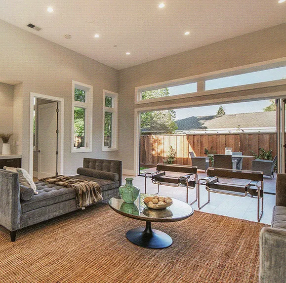 A modern living room with a large, open-plan design featuring a gray sofa, a round glass coffee table, and large sliding glass doors leading to a patio with outdoor furniture.